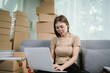 © Kieng - A young Asian woman sits on a sofa, writing a message on a package for delivery, surrounded by boxes and a laptop. She is focused on managing her online shopping orders.