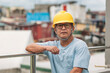 © Mdv Edwards - Mature Southeast Asian man in hardhat poses on balcony with serious expression, gazing over squatter area during work break.