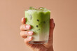 © Penkinn - A hand holding a glass of iced matcha latte, with separated green tea, milk layers. Visible ice cubes. commercial drink. on pastel beige background.