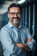 © Nadzeya - A friendly and confident middle-aged system administrator with a beard and glasses smiles at the camera. Standing with arms crossed in a data center, he projects professionalism and expertise