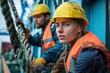 © Nadzeya - Two diverse deckhands, a male and female, wearing yellow hard hats and orange safety vests, work with thick ropes on a ship. Their focused expressions show their determination