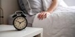 © Risat Designer - Close up of a vintage alarm clock on a nightstand next to a person waking up in a cozy bed with white linens