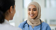 © Mono - A smiling female healthcare professional wearing a hijab and scrubs speaks with a colleague in a bright hospital setting.
