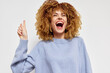 © SHOTPRIME STUDIO - Happy woman with curly hair smiling and pointing upwards, wearing a light blue sweater against a neutral background Joyful expression showcasing excitement and positivity