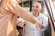 © Pixel-Shot - Young caregiver helping senior woman with stick to stand up from car outdoors