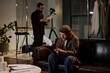 © TrueFrame Collective - Caucasian young adult man sitting on sofa reading script during acting audition while middle aged Caucasian man standing in background holding papers near camera equipment