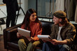 © TrueFrame Collective - Young adult Caucasian man and young adult Hispanic woman sitting on sofa holding scripts, engaging in conversation during acting audition, professional studio equipment visible in background
