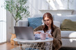 © Jirakul - woman working on her laptop at home, focused on her tasks while sitting on floor. cozy atmosphere with natural light and plants enhances scene