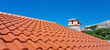 © nahhan - A terracotta tiled roof with a stone chimney under a clear blue sky, with a mountainous landscape in the background