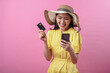 © Daniel - Asian woman wearing a summer hat and yellow dress smiling while holding a credit card and smartphone, standing in front of a pink background