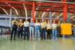 © AU USAnakul - Diverse team of factory workers, engineers, and office staff pose confidently inside modern steel roofing sheet production plant. Their stance reflects unity, pride, and industrial teamwork