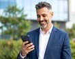 © aziz - Happy stylish older busy businessman investor holding smartphone looking at cellphone doing banking payments. Middle aged business man using mobile cell phone working outdoors. Candid photo.