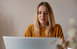 © My Ocean studio - Young woman in a mustard sweater working on a laptop at home. Focused expression, casual setting, soft daylight, and neutral background. Remote work or online learning concept.