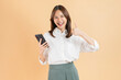 © sitthiphong - Cheerful beautiful Asian business woman holding smartphone and shows like sign on beige color background