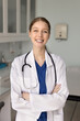 © fizkes - Happy beautiful young physician woman in lab coat posing for profile picture in doctor office, standing with hands folded in confident gesture, looking at camera with toothy smile. Vertical portrait