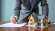 © Soyaeb - Businessman in a suit signing a real estate contract with a model house on the desk, representing a property purchase or mortgage deal.