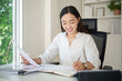 © matinee - Asian businesswoman reviewing documents and taking notes at a bright office desk.
