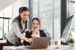 © kenchiro168 - Two Asian businesswomen in formal attire working together at modern office desk, one standing and pointing at laptop screen while other sits and smiles, teamwork and collaboration