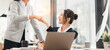 © kenchiro168 - Two businesswomen shaking hands in bright modern office, smiling and celebrating success with laptop and coffee cups on desk, showing teamwork and positive collaboration