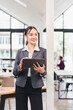 © kenchiro168 - Young businesswoman holding digital tablet standing near pillar in modern office with natural light, looking away with confident smile and professional attire