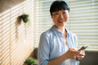 © Marko Geber - Smiling Asian woman holding smartphone while standing in sunlit modern office