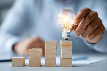 Poster - Hand Placing a Glowing Light Bulb on Stacked Wooden Blocks Representing Growth and Innovation in a Business Setting