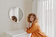 © SHOTPRIME STUDIO - Smiling young woman with curly hair in orange blouse leaning on a white dresser, reflecting in a round mirror, relaxed indoor setting highlighting positivity and style