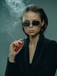 © SHOTPRIME STUDIO - Stylish woman vaping in sunglasses with smoke, wearing black outfit, confident and modern lifestyle concept.