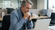 © MR RIHAD - A middleaged businessman with gray hair, wearing a blue jacket, is thoughtfully focused on his laptop screen while sitting at his desk in a contemporary office setting