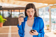© luismolinero - Young pretty woman at outdoors using mobile phone and holding a coffee with surprised expression