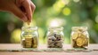 © Cherry - Hand adding coin to one of three glass jars filled with various coins, sitting on wooden surface against blurred green background