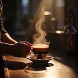 Hand holding a transparent cup of steaming espresso lungo indoors, capturing rich coffee color and rising steam above a wooden table in warm light.
