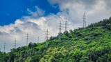 Power lines on a green hill with dense vegetation and cloudy sky. Concept of energy, infrastructure and power transmission.