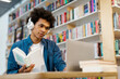 © Home-stock - Focused black male college student wearing headphone, sitting at desk in library, studying, holding book and using laptop for research or project