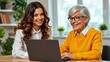 © IgorImageDoc - two women sitting desk office woman young woman long brown hair wearing white blouse smiling camera she sitting next older woman who wearing yellow sweater glasses both looking laptop screen appear
