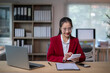 © Apichat - woman in a red suit is sitting at a desk with a laptop and a tablet