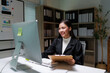 © Crystal - Young happy Asian businesswoman working with desktop computer and holding clipboard, smiling and looking at computer screen in modern office in the evening