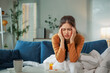 © Crystal - Young woman suffering from strong headache while sitting on sofa at home with medicines and hot drink on table, she touching temples with fingers, feeling unhealthy