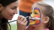 © MdMohiuddin - Young Girl Smiles During Face Painting Session with Artistic Woman