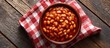 © rich - A bowl of baked beans sits atop a red-checkered napkin on a rustic wooden surface