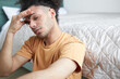 © Anatoliy Karlyuk - Young man in orange shirt holding his forehead with closed eyes, suffering migraine or headache, sitting on floor in his bedroom with closed eyes, feeling unwell after waking and getting up