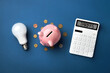 © PixelArt_25 - A pink piggy bank with coins, a calculator displaying 'TARIFFS', and a light bulb on a blue background, symbolizing energy cost increases due to tariffs.Top view, flat lay.