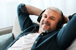 © Prostock-studio - Happy mature man reclining on couch at home, listening to music and smiling at camera, using wireless modern headset, closeup, copy space. Modern technologies and entertainment concept
