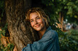 © Audrey - Young woman hugs tree in park. Smiling face, sunlight, nature background. Blonde hair, denim shirt. Joyful, happy moments outdoors, feeling good. Woman enjoys fresh air, forest.