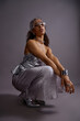 © Seventyfour - Vertical portrait of young adult multiethnic woman crouching in studio, wearing metallic accessories and futuristic outfit, looking confidently into camera during fashion photoshoot session