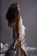© Seventyfour - Vertical shot of young multiethnic woman standing in profile wearing metallic fashion outfit, holding silver and gold fabric, curly hair adorned with decorative headpiece during fashion photoshoot