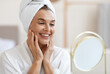 © Prostock-studio - Beautiful middle aged woman in white bathrobe and with turban on her head looking at mirror, touching her face skin and smiling, using best anti-aging cosmetics, home interior, copy space