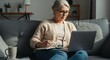 © PepeCura - Focused Senior Woman Working on Laptop and Writing in a Notebook on a Couch