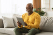 © Prostock-studio - Mature African American Man Using Digital Tablet Sitting On Sofa At Home. Senior Male Browsing Internet On Computer Reading And Scrolling News Feed Online. People And Gadgets