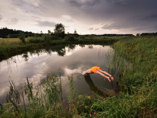 Wall Mural - a man is jumping into a pond wearing an orange swimsuit at dusk. the sky is cloudy and the water is 